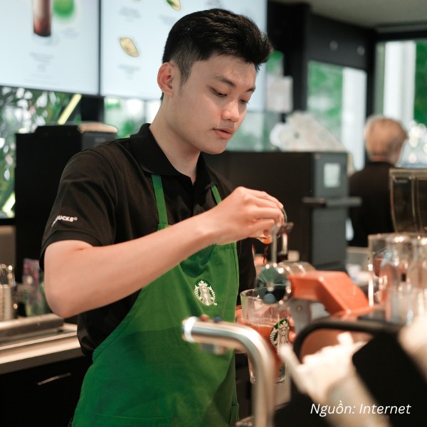 Nhân viên Starbucks chuyên nghiệp với đồng phục polo đen phối tạp dề xanh lá đặc trưng.